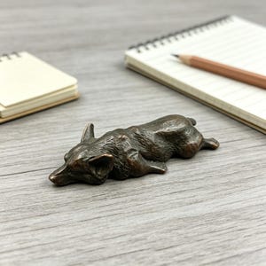 May include: A small, dark bronze-colored metal figurine of a dog lying down in a resting position. The sculpture features detailed textures representing fur and the distinct pointed ears of a corgi, captured in a solid, cast metal form.