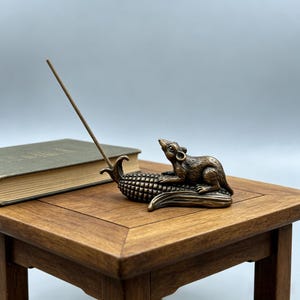 May include: A bronze-colored incense holder featuring a rat resting on a corn cob. An incense stick is inserted into the holder. The holder sits on a wooden table next to a book with a green cover.