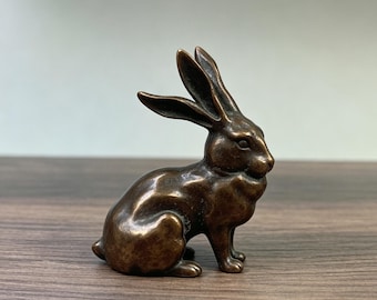 May include: A bronze-colored rabbit figurine in a seated position. The sculpture has detailed features, including long ears and a textured surface. The rabbit is a decorative item, suitable for home decor or as a collectible. The figurine is approximately 4 inches tall.