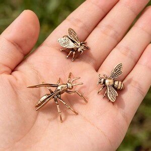 May include: Three small metallic insect figurines in a bronze tone. One figurine depicts a wasp with elongated wings and legs, while the other two are smaller bee-shaped ornaments featuring detailed wing patterns and segmented bodies.