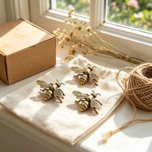 May include: Three antique brass-toned metal bee ornaments featuring detailed wings and striped bodies. The bees are arranged on a light-colored fabric pouch, accompanied by a brown cardboard box and a ball of natural jute twine nearby.