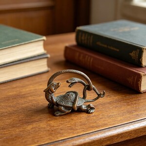 Puede incluir: Una pequeña figura de metal en tono bronce que representa una tortuga con caparazón texturizado y una serpiente enroscada alrededor de su cuerpo. La cabeza de la serpiente está elevada frente a la tortuga, con escamas detalladas en ambas.