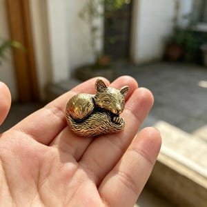 May include: A small, metallic gold-colored figurine of a curled-up fox. The fox is depicted in a resting position with its tail wrapped around its body, featuring detailed engraved lines that mimic the texture of fur across its entire surface.