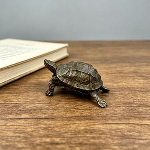 May include: A bronze-colored turtle figurine with detailed shell patterns and textured skin. The turtle is posed on a wooden surface, with a book in the background. The figurine is a decorative item.