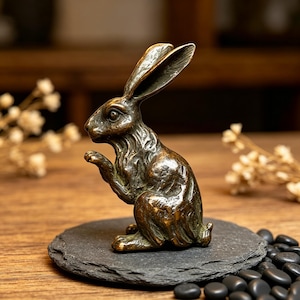 May include: A bronze-toned metal figurine of a rabbit in a seated, alert posture with long ears and textured fur details. The sculpture is positioned on a circular, dark grey slate base, showcasing a weathered, metallic finish throughout the piece.