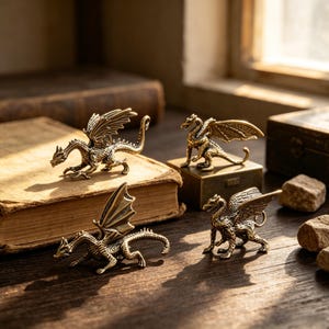 May include: Four small, detailed brass dragon figurines with textured scales and spread wings. One dragon stands on a small rectangular brass box, while another rests on an old book. The figures feature intricate carvings and a metallic finish.