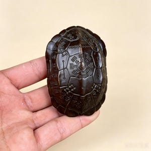 May include: A dark brown, carved wooden turtle shell ornament featuring intricate engravings of traditional Chinese trigrams and characters across its surface. The shell has a segmented, rounded design with detailed line work etched into the wood.
