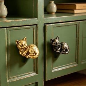 Può includere: Due pomelli per armadietto a forma di gatto fissati su un mobile in legno verde. Un pomello è in metallo color oro lucido, mentre l'altro è in metallo color bronzo antico più scuro. Entrambi i pomelli presentano una figura felina in riposo.