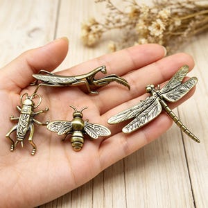 Könnte beinhalten: Ein Set aus vier Insektenfiguren aus antikem messingfarbenem Metall, bestehend aus einer Gottesanbeterin, einer Heuschrecke, einer Biene und einer Libelle. Jedes Insekt weist detaillierte geätzte Flügel- und Körperstrukturen mit dunklen Akzenten auf.