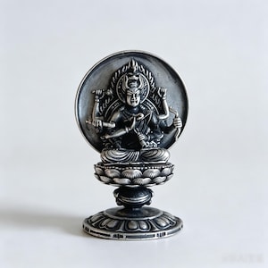 May include: A metallic silver-toned figurine of a multi-armed deity seated in a meditative pose upon a lotus pedestal. The figure is framed by a circular halo and features intricate detailing on the arms, face, and the layered lotus base.