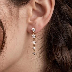 Puede incluir: Un pendiente colgante de tono dorado que presenta una cadena vertical de cinco piedras pequeñas, claras y de corte redondo. El pendiente se sujeta al lóbulo con un perno, creando una apariencia lineal, delicada y colgante.