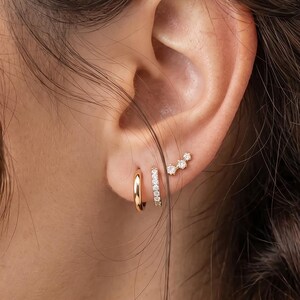 Peut inclure: Un ensemble de trois boucles d'oreilles dorées portées au lobe et au cartilage. La première est un anneau doré poli, la deuxième est un anneau fin serti de petites pierres claires, et la troisième est un petit clou doré à trois pierres.