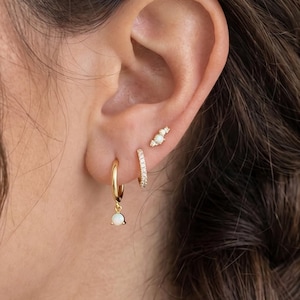 Könnte beinhalten: Ein Set aus drei goldenen Ohrringen, die am Ohrläppchen und am Knorpel getragen werden. Der unterste Ohrring ist ein goldener Creolen-Ring mit einem kleinen weißen Opal-Anhänger. Darüber befindet sich ein dünner Goldring und ein kleiner Goldstecker.