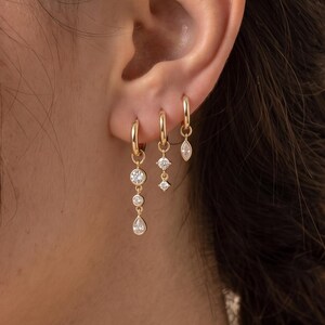 May include: Three gold hoop earrings worn in a single earlobe. The first hoop features a long drop with two round clear stones and a teardrop-shaped stone. The second hoop has two small round clear stones. The third hoop has a marquise-cut stone.