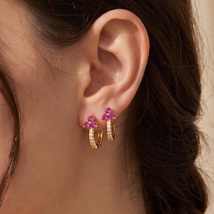 May include: A pair of gold hoop earrings featuring a small cluster of three vibrant pink stones at the top. The hoops are adorned with a row of tiny clear stones along the front edge, creating a delicate and refined look for the earlobe.