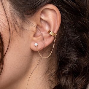 May include: A gold-toned ear cuff featuring three parallel bands connected by a delicate chain to a single stud earring with a clear, round-cut stone. The jewelry is worn on the ear, with the cuff positioned on the upper cartilage and the stud.