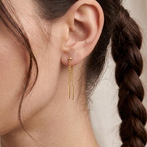 May include: A gold-toned earring featuring a small spherical stud connected to two delicate chains of varying lengths, each ending in a slender, polished cylindrical bar pendant that hangs vertically from the earlobe.