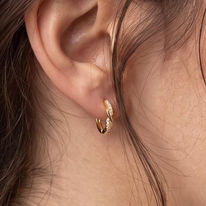 May include: A small gold hoop earring featuring a twisted design with embedded clear stones. The earring is worn in the earlobe, showcasing a metallic finish and a circular shape that curves around the lower part of the ear cartilage.