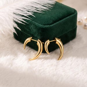 May include: A pair of gold-toned earrings featuring a curved, tapered horn or tusk shape. The earrings have a smooth, polished metallic finish and a post-back closure mechanism, designed to wrap around the earlobe with a sleek, pointed aesthetic.
