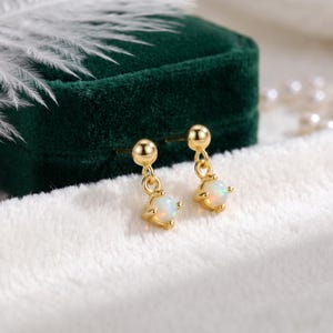 May include: A pair of gold drop earrings featuring a small round opal stone suspended from a gold ball stud. The opal stones exhibit a subtle iridescent play of color, and the metal components have a polished gold finish, creating a refined look.