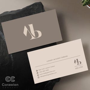 Op de afbeelding: Twee rechthoekige visitekaartjes in een taupe en crème kleurenschema. Eén kaart heeft een groot gestileerd AB-monogram. De andere kaart toont de tekst LUXURY IN EVERY THREAD, het AB-monogram, ABLURE en contactgegevens voor New York, Londen en Dubai.