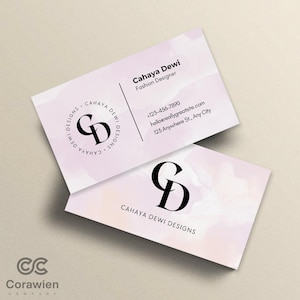 Pode incluir: Dois cartões de visita retangulares com um fundo de aguarela rosa pálido. Um cartão exibe o nome Cahaya Dewi, Fashion Designer, detalhes de contacto e um logótipo circular com as letras CD. O outro cartão apresenta um grande logótipo CD preto.