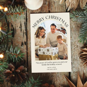 Puede incluir: Una tarjeta de Navidad con una foto de una familia de tres abriendo un regalo. La tarjeta tiene un arco dorado con el texto "Merry Christmas" y el a&ntilde;o "2024". El texto "May peace of God be upon you and your family this holiday season! With love, The Miller's" est&aacute; impreso debajo de la foto.