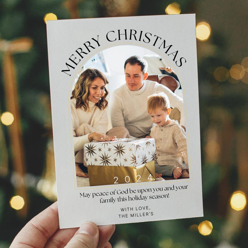 Puede incluir: Una tarjeta de Navidad con una foto de una familia de tres. La tarjeta tiene un arco dorado con el texto "Merry Christmas" en negro. La foto muestra una familia de tres, una mujer, un hombre y un ni&ntilde;o peque&ntilde;o, de pie frente a un regalo envuelto. El a&ntilde;o 2024 est&aacute; impreso en dorado debajo de la foto. El texto "May peace of God be upon you and your family this holiday season!" est&aacute; impreso debajo del a&ntilde;o. El texto "With love, The Miller's" est&aacute; impreso en la parte inferior de la tarjeta.