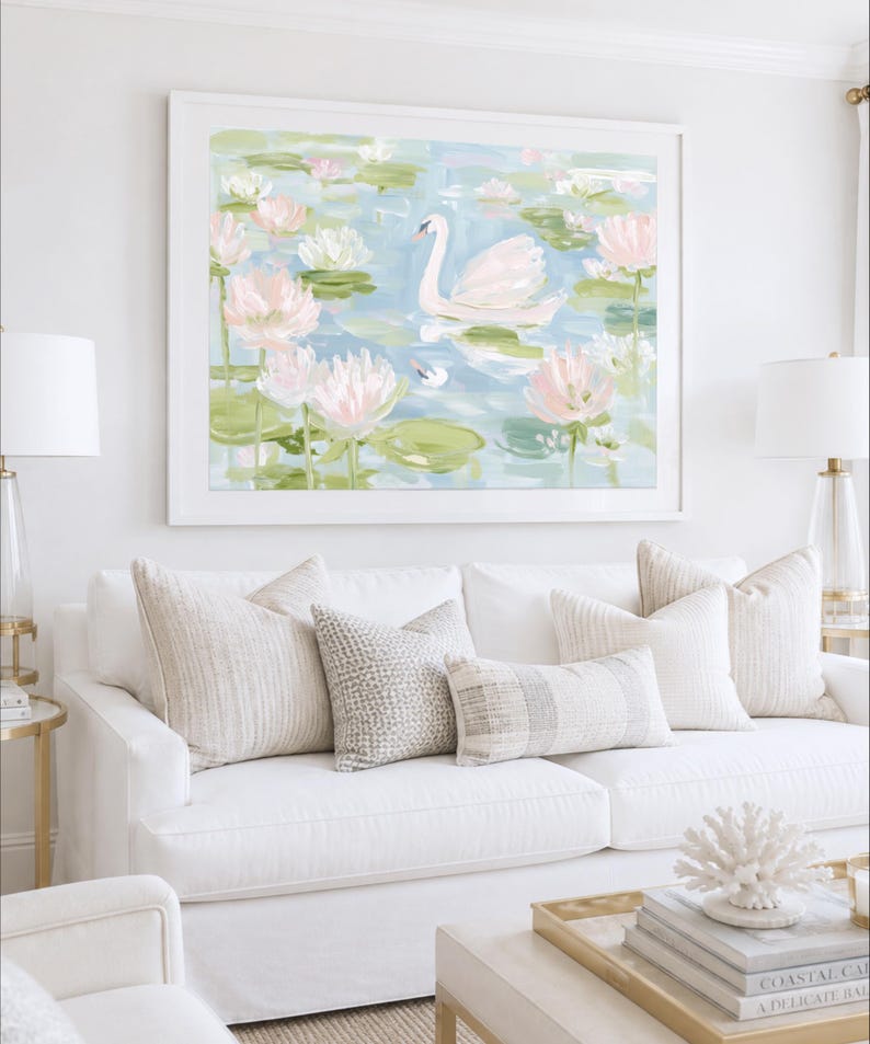 May include: A framed wall art print featuring an impressionistic painting of a white swan swimming in a pond filled with light pink and white water lilies, green lily pads, and soft blue water, displayed in a white frame above a white sofa.