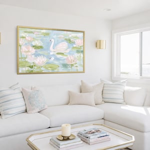 May include: A rectangular wall art print in a gold frame featuring a painting of a white swan swimming among pink lotus flowers and green lily pads on a light blue water surface, displayed above a white sofa with decorative throw pillows.