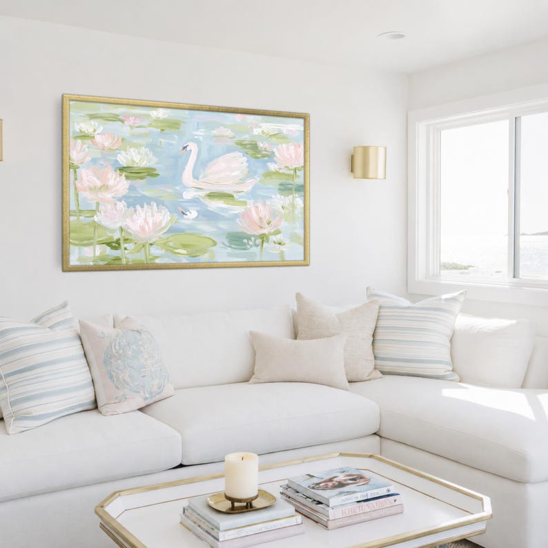 May include: A rectangular wall art print in a gold frame featuring a painting of a white swan swimming among pink lotus flowers and green lily pads on a light blue water surface, displayed above a white sofa with decorative throw pillows.