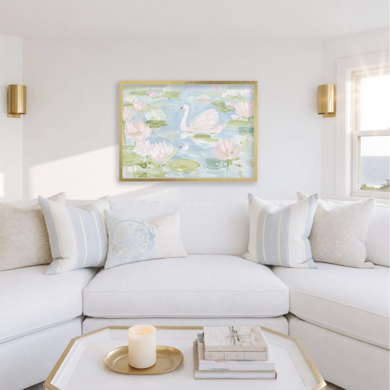 May include: A rectangular wall art print in a gold frame featuring an impressionistic painting of a white swan swimming among pink lotus flowers and green lily pads on a light blue water surface, displayed above a white sectional sofa.