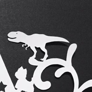 Puede incluir: Un recorte de papel blanco de un dinosaurio Tyrannosaurus Rex se alza sobre un remolino decorativo. La imagen est&aacute; sobre un fondo negro. El dinosaurio tiene una boca sonriente y est&aacute; en posici&oacute;n de caminar. El dise&ntilde;o incluye formas de estrella.