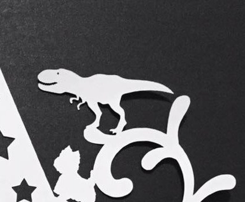 Puede incluir: Un recorte de papel blanco de un dinosaurio Tyrannosaurus Rex se alza sobre un remolino decorativo. La imagen est&aacute; sobre un fondo negro. El dinosaurio tiene una boca sonriente y est&aacute; en posici&oacute;n de caminar. El dise&ntilde;o incluye formas de estrella.