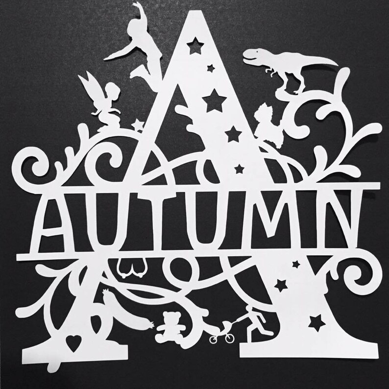 Puede incluir: Recorte de papel blanco con el nombre "AUTUMN", con la letra "A" como elemento principal. La letra est&aacute; adornada con estrellas, un hada, un dinosaurio, un ni&ntilde;o y otros motivos l&uacute;dicos. El fondo es negro.