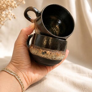 以下が含まれることがあります： 光沢のある黒い内側と上部外側が特徴の積み重ねられた2つの陶器製マグカップ。中央には質感のある多孔質のベージュと茶色の帯があり、底は滑らかな黒色です。各マグカップの側面には小さな湾曲したハンドルが付いています。