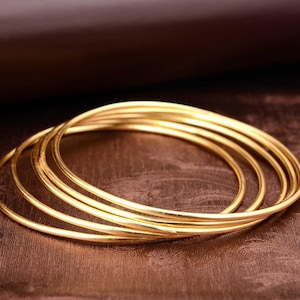 Puede incluir: Un conjunto de cinco brazaletes circulares y delgados de oro con un acabado liso y pulido. Las pulseras están apiladas superponiéndose ligeramente entre sí, creando una apariencia en capas con un color amarillo metálico uniforme.