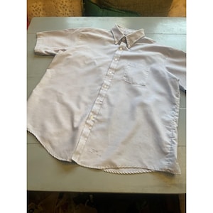 May include: A light blue short-sleeved button-down shirt with a button-down collar and a chest pocket. The shirt has a relaxed fit and a curved hemline. There is text printed on the lower left side of the shirt.