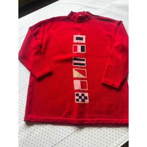May include: A bright red long-sleeve mock neck sweater featuring a vertical column of six distinct nautical signal flags centered on the front. The flags are embroidered in various colors including black, white, pink, and tan, arranged in a single line.