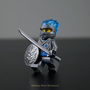 Könnte beinhalten: Eine Lego-Minifigur eines Ninja-Kriegers, gekleidet in grauer und blauer Rüstung, hält ein silbernes Schwert und einen Schild. Die Figur hat ein blaues Haarteil und eine schwarze Maske. Der Schild hat ein detailliertes silbernes Design. Der Text "Building Block Minifigures" steht unten.