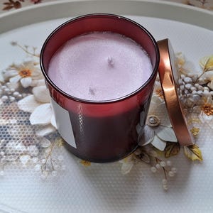 Puede incluir: Una vela cilíndrica en un frasco de vidrio rojo oscuro con una superficie de cera color lavanda claro y dos mechas. Una tapa metálica de color cobre descansa junto al frasco. La vela presenta una etiqueta rectangular blanca lisa.