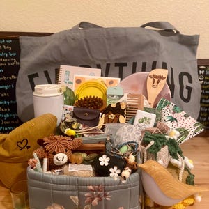 Puede incluir: Una colección de artículos que incluye una bolsa de tela gris con la palabra EVERYTHING, una cesta de almacenamiento acolchada con flores, una gorra de béisbol color mostaza con un corazón, una espátula de madera y varios accesorios.
