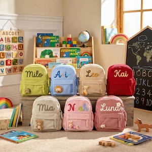 May include: Seven corduroy backpacks in various colors including lime green, blue, beige, red, cream, pink, and dusty rose. Each bag features a name embroidered in cursive script and a small plush animal attached to the front zippered pocket.