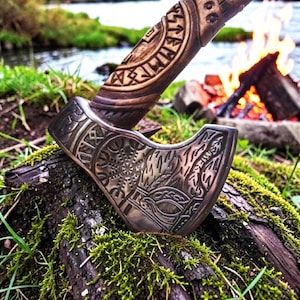 May include: A metal axe head with intricate engravings of a Vegvisir symbol and knotwork patterns, attached to a dark wooden handle featuring carved runic inscriptions. The axe is embedded into a mossy log, showcasing detailed metalwork.