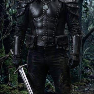 May include: A dark leather chest plate with silver studs, matching shoulder pauldrons, and forearm bracers. The outfit includes a belt with two attached pouches, dark trousers, and tall boots. A silver medallion hangs from the neck, and a long steel sword is held in the right hand.