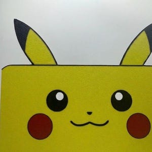 May include: A yellow Pikachu-themed box with black-tipped ears, black eyes, red cheeks, and a smiling mouth. The box is a bright yellow color and has a simple, cartoonish design.