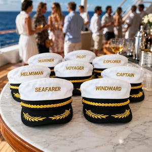 May include: A collection of white and black nautical caps with gold braided trim and embroidered gold laurel leaves on the brim. Each cap features a unique embroidered word on the front, including Mariner, Pilot, Explorer, Helmsman, Voyager, Captain, Seafarer, and Windward.