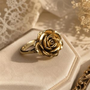 Puede incluir: Un anillo de tono dorado que presenta una flor de rosa tridimensional como pieza central. Los pétalos de metal están superpuestos y esculpidos para imitar la forma natural de una flor, unidos a una banda lisa y delgada.