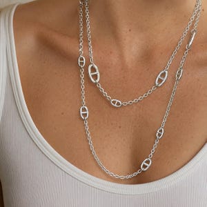 Könnte beinhalten: Eine lange silberne Halskette mit einer Reihe gleichmäßig verteilter ovaler Glieder, die in der Mitte einen horizontalen Steg aufweisen und mit runden Standardkettengliedern abwechseln. Die Kette wird in einem zweilagigen Stil getragen.