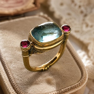 May include: A gold ring featuring a large, rectangular, faceted light blue gemstone set in the center, flanked by two smaller, round, deep red gemstones on either side. The metal band has a textured, aged finish with visible decorative detailing.